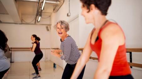 Zwei Frauen nehmen an einem Fitnesskurs teil und lachen dabei. Im Hintergrund sieht man weitere Teilnehmer.